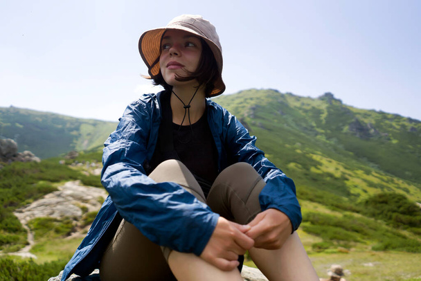 帽子とウィンドブレーカーの幸せな若い女の子は岩の上に座り,彼女の旅の間に山,丘,森で素晴らしい景色を楽しんで,野生でハイキングしています. 女性が活発なライフスタイルを過ごす. - 写真・画像