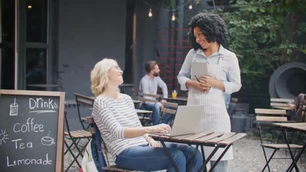 Friendly African American waitress holding clipboard while receiving client on cafe terrace. Cheerful female customer making order while sitting at table in sunny day. People communicating outdoors. - Footage, Video