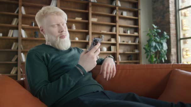 Close up view of serious middle aged man with blond hair sitting on leather sofa using phone in modern urban loft apartment, texting message scrolling news. Technology lifestyle slow motion dolly shot - Кадры, видео