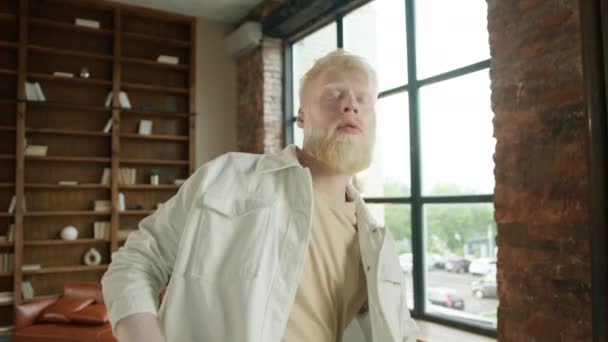 Echte mensen dansen in slow motion 4K. Jonge expressieve blonde man met baard die vogue danst in loft studio met creatief interieur, grote ramen en bibliotheek op houten muur. Vreemde albino jongen. - Video