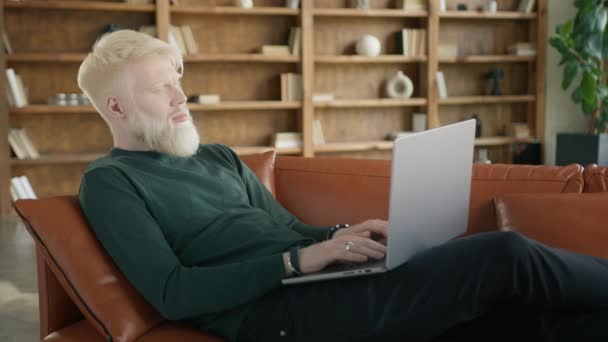 Empresario albino rubio concentrado que trabaja a distancia escribiendo en el portátil en el apartamento loft moderno 4K lento. Enfocado chico millennial serio freelancer utilizando el ordenador portátil en el sofá en casa sala de estar de la oficina - Imágenes, Vídeo
