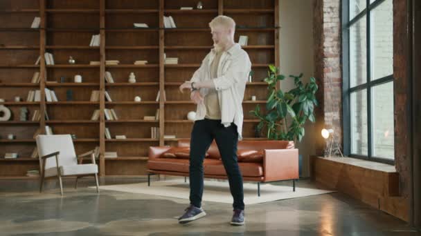 Happy young man dancing in loft style living room. Man wearing hipster casual clothes, jumping and moving hands around in ecstatic moves. Blond bearded guy listening to music at home on sunny morning - Footage, Video