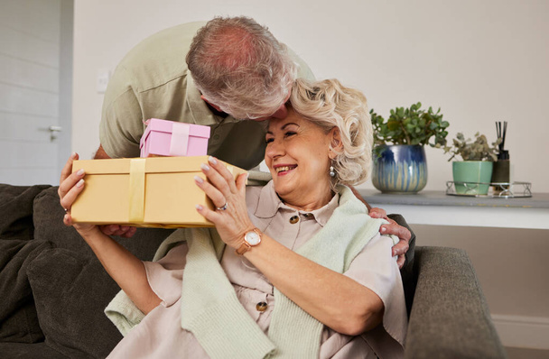 Gift, surprise and a senior couple on their anniversary for celebration in their home living room together. Love, present and birthday with a husband giving his wife a box while sitting on a sofa. - Foto, immagini