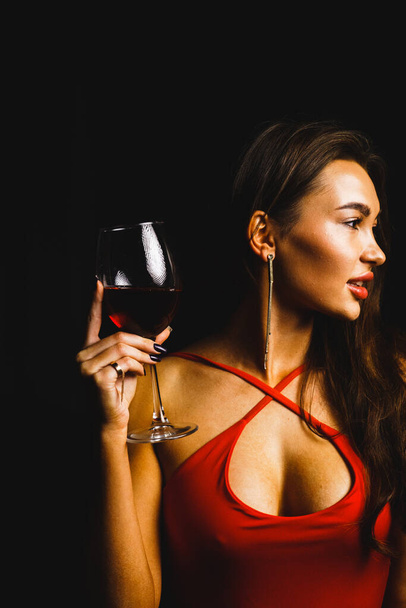 Ένα ποτήρι κόκκινο κρασί στα χέρια μιας σέξι γυναίκας με κόκκινο φόρεμα. - Φωτογραφία, εικόνα