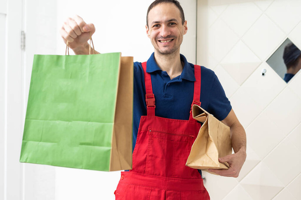 Obrázek šťastného mladého doručovatele v červené čepici stojícího s poštovní schránkou izolovanou nad bílým pozadím. Kvalitní fotografie - Fotografie, Obrázek