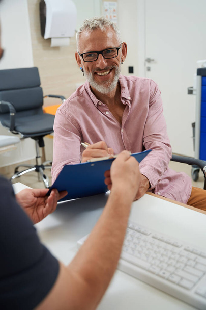 Parrakas iäkäs mies allekirjoittaa asiakirjoja lääkäreiden toimistossa, miehet istuvat toimistopöydässä - Valokuva, kuva