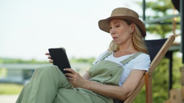 Uma mulher de meia-idade passa férias de verão no campo, desfruta do ar livre e relaxa após o trabalho no jardim, sentado em uma reclinável. Uma senhora lê histórias engraçadas ou anedotas, usando um tablet - Filmagem, Vídeo