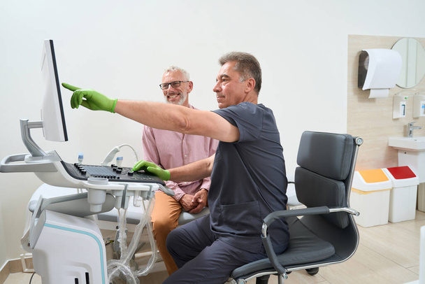 Deneyimli uzolog, ultrason odasında gri saçlı bir hastaya danışıyor. Klinik modern ekipman kullanıyor. - Fotoğraf, Görsel