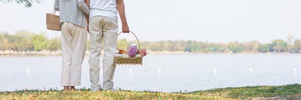Nuori pari kuljettaa piknik kori ja syleilee yhdessä seisoessaan lähellä järven puistossa. - Valokuva, kuva