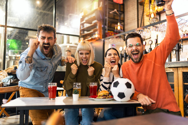 Emocionados aficionados al fútbol celebrando mientras ven el partido de fútbol en la televisión en el bar. - Foto, Imagen