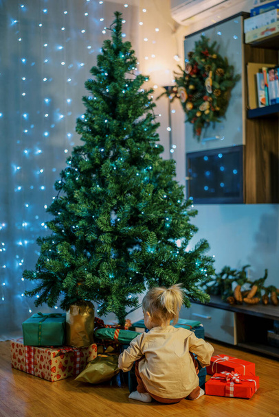 A kislány a karácsonyfa alatt ül a padlón, és ajándékokat néz. Visszanézni. Kiváló minőségű fénykép - Fotó, kép
