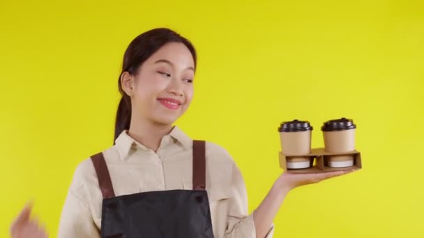 Retrato joven barista asiática mujer usando delantal sosteniendo taza de café y presentando sobre fondo amarillo, camarera alegre y mostrando y pulgares hacia arriba, pequeña empresa o startup, camarero de cafetería. - Imágenes, Vídeo
