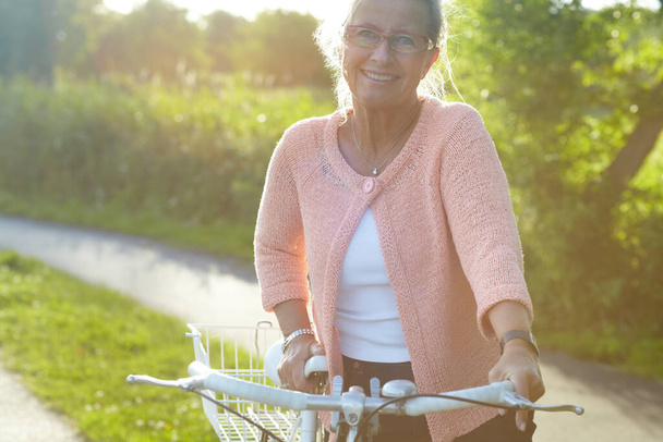 Retrato, sonrisa y anciana en bicicleta al aire libre, ejercicio y fitness en verano. Feliz, persona mayor en bicicleta en la naturaleza o jardín para el ciclismo en el campo para la salud corporal, viajes y gafas. - Foto, imagen