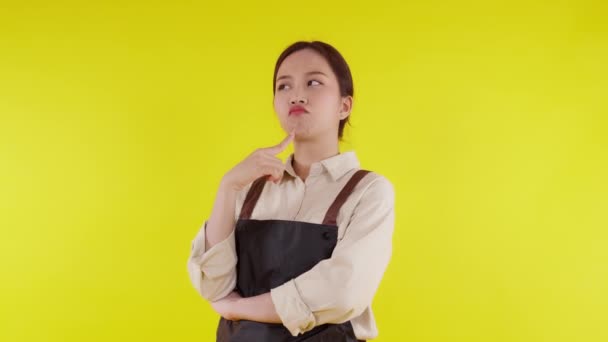 Retrato de una joven barista asiática con un gesto de delantal con una idea de pensamiento y éxito sobre fondo amarillo, camarera o emprendedora pensada y emocionada, pequeña empresa o startup, camarera de cafetería. - Metraje, vídeo