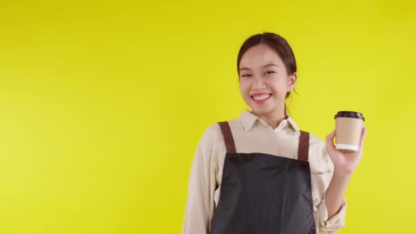 Portrét mladé asijské barista žena na zástěře drží šálek kávy a prezentuje na žlutém pozadí, servírka nebo podnikatel veselý a show, malý obchod nebo start-up, číšník kavárny. - Záběry, video
