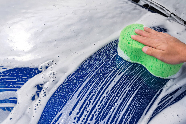 An washing a soapy blue car with a green sponge. - Foto, afbeelding