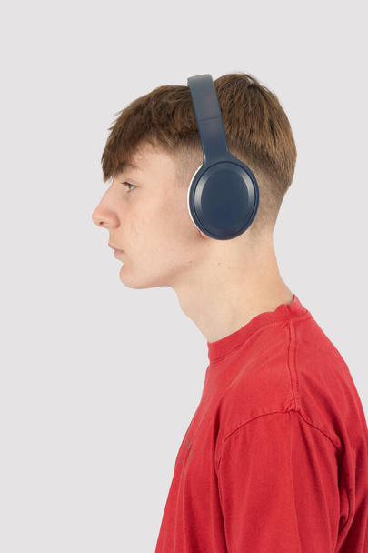 Biały 15-letni chłopiec, ubrany w czerwony t-shirt, słuchający muzyki przez słuchawki patrzące z boku - Zdjęcie, obraz