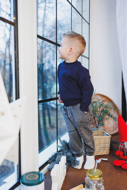 Un niño de 2 años se sienta junto a la ventana en el Año Nuevo. Ambiente de Año Nuevo en casa. El niño está esperando el año nuevo. - Foto, imagen