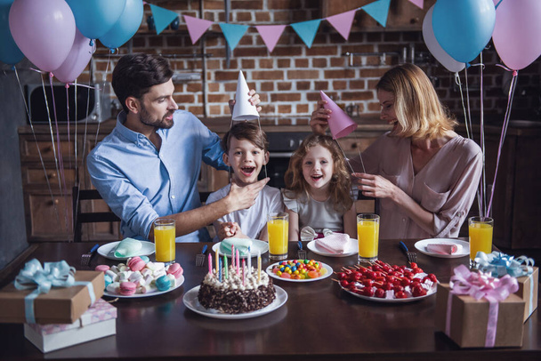 Happy family is smiling while sitting at the table in decorated kitchen during birthday celebration, kids are looking at camera - Photo, Image