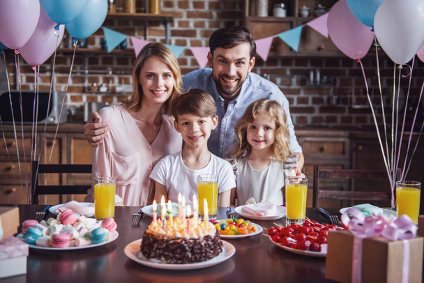 Boldog család keresi a kamera, és mosolyogva, miközben ül az asztalnál, berendezett konyha, születésnapi ünnepség során portréja - Fotó, kép