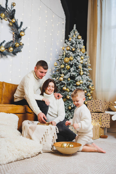 Una alegre familia joven con un niño pequeño descansa cerca del árbol de Navidad en casa. Pareja familiar con decoración navideña en casa. Navidad, Año Nuevo - es hora de celebrar - Foto, Imagen