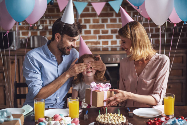 glückliche Familie sitzt bei Geburtstagsfeier am Tisch in dekorierter Küche, Papa bedeckt Tochter die Augen, während Mama ein Geschenk in der Hand hält - Foto, Bild