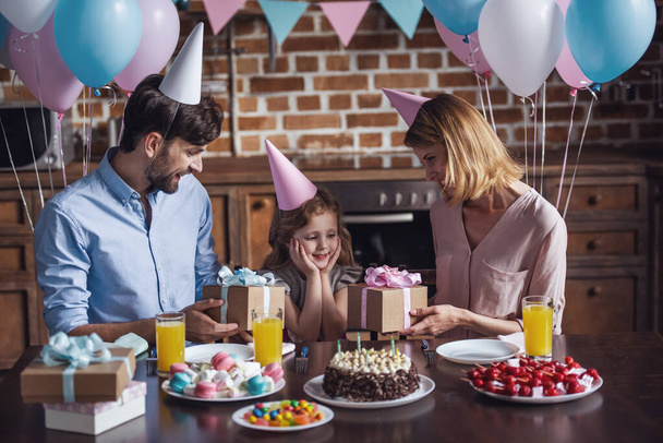 Glückliche Eltern beschenken ihre Tochter, während sie bei der Geburtstagsfeier am Tisch in der dekorierten Küche sitzen - Foto, Bild