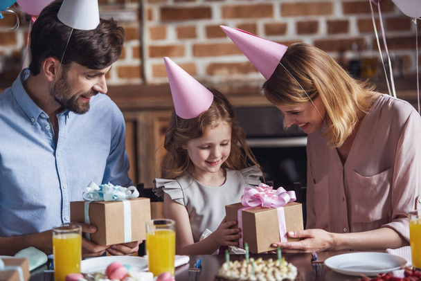 Los padres felices están dando regalos a su hija mientras están sentados en la mesa en la cocina decorada durante la celebración del cumpleaños
 - Foto, imagen