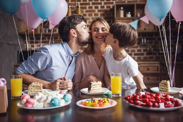 Familie feiert Muttergeburtstag in dekorierter Küche. schöne Frau blickt in die Kamera und lächelt, während Mann und Sohn sie auf die Wangen küssen - Foto, Bild
