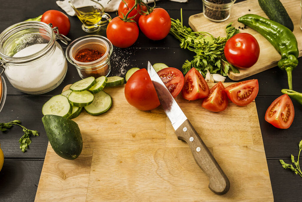   Stilleven met wat rijpe gesplitste tomaten samen met knoflook, komkommerschijfjes, paprika, een fles zout, hele citroenen, olijfolie en een mes op een houten plank - Foto, afbeelding