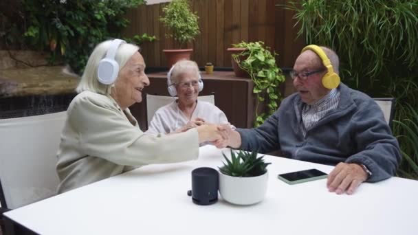 Video de ancianos estrechando la mano mientras escuchan un podcast con auriculares en un hogar de ancianos - Metraje, vídeo