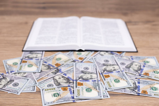 Открытая книга Библии на столе. Банкноты по доллару. Выбор: богатство или Бог. Богатым трудно войти в Царствие Божие - Фото, изображение
