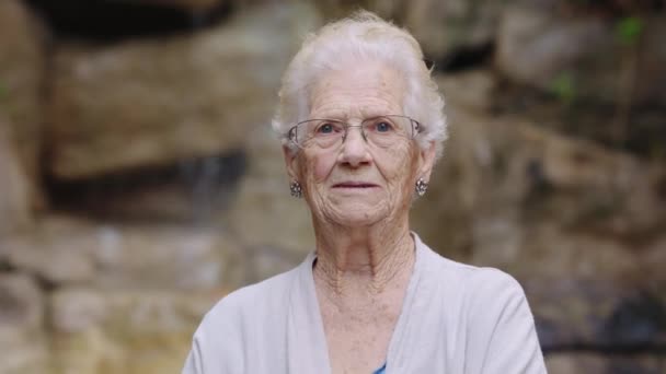 Videó egy idős nőről, aki mosolyog és integet a kamerának egy nővérház kertjében. - Felvétel, videó