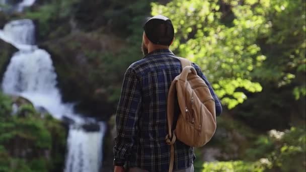 hombre admira un paisaje de montaña y una cascada durante una caminata - Metraje, vídeo