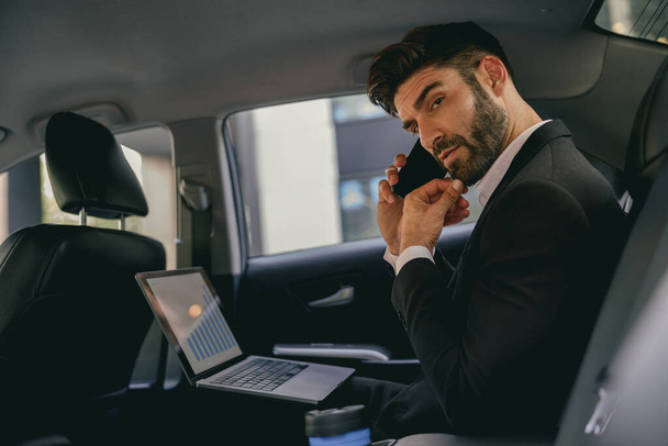 Hombre de negocios guapo en traje usando el ordenador personal y haciendo una llamada telefónica en taxi - Foto, Imagen