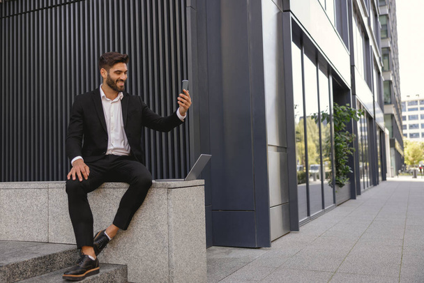 Empresário sorridente faz uma selfie no celular sentado no banco perto do prédio de escritórios - Foto, Imagem