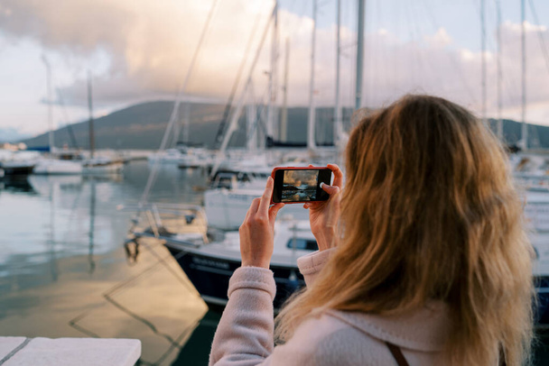 Kız, dağların eteklerinde yatlı bir marinanın akıllı telefonuyla fotoğraflarını çekiyor. Arkadan bak. Yüksek kalite fotoğraf - Fotoğraf, Görsel
