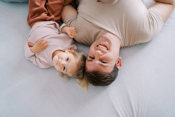 Papá sonriente con su hijita acostada en la cama, tocando las tapas. Vista superior. Foto de alta calidad - Foto, Imagen