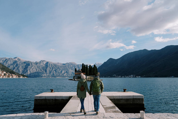 Pari kävelyä käsi kädessä laiturilla, josta on näköala St. George Islandille. Montenegrossa. Taustakuva. Laadukas kuva - Valokuva, kuva