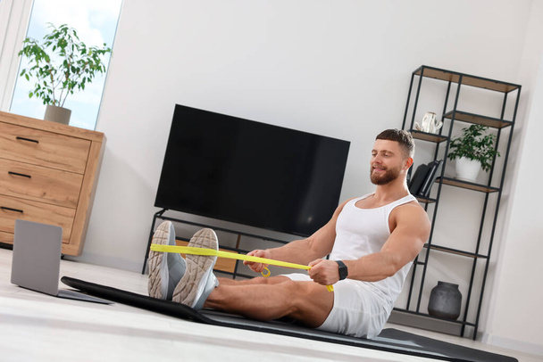 Muscular man doing exercise with elastic resistance band near laptop on mat at home - Foto, immagini