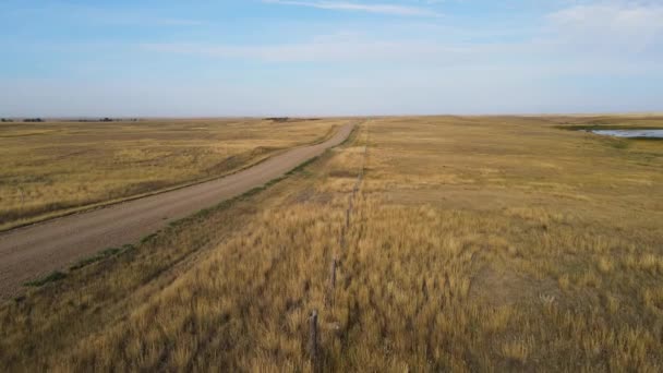 Drone vliegt laag boven een droog West-Amerikaans landschap. Het pad van de drone volgt een prikkeldraad hek naast een grindweg. De blauwe lucht heeft dunne witte wolken. - Video