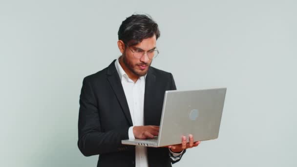 Взволнованный счастливый индийский бизнесмен печатает на ноутбуке, работает над проектом, наслаждается результатами, выигрывает в лотерею, празднует успех, онлайн-покупки. Человек в формальном костюме изолирован на сером фоне - Кадры, видео