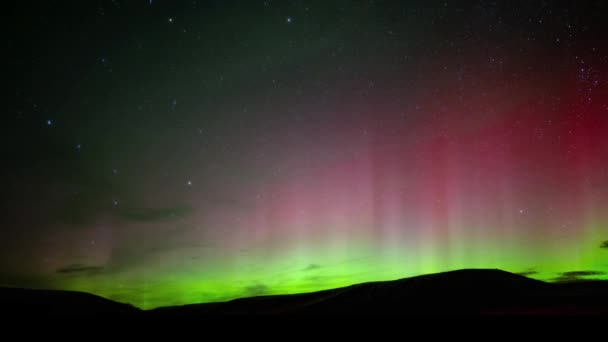 Bright green and red aurora flash in a star filled sky above rolling hills. The Big Dipper can be seen on the left side and the stars are moving slowly. - Footage, Video