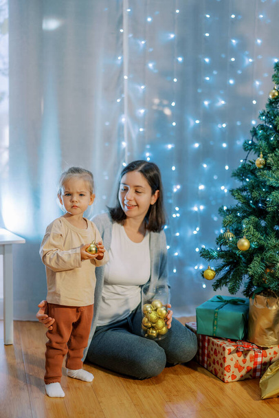 Niña con una pelota en las manos está cerca de su madre sentada cerca del árbol de Navidad con una caja de decoraciones navideñas. Foto de alta calidad - Foto, imagen