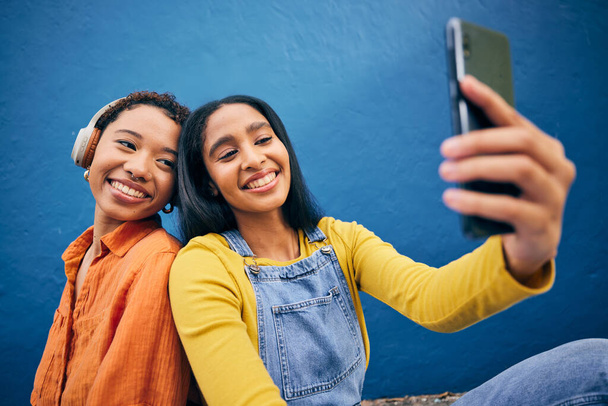Phone, selfie and women in city on blue background online for social media, internet and profile picture. Friendship, happy and female people relax on smartphone listening to song, track and radio. - Photo, Image