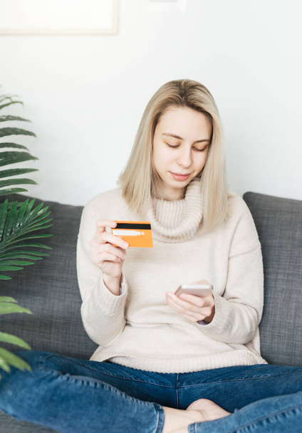 Young woman holding smartphone and banking credit card, involved in online mobile shopping at home, happy female shopper purchasing goods or services in internet store. - Photo, Image