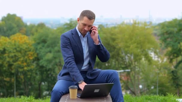 Κάμερα πλησιάζει νεαρός όμορφος επιχειρηματίας κάθεται σε ένα παγκάκι κουβεντιάζοντας στο smartphone και χρησιμοποιώντας φορητό υπολογιστή, ενώ πίνοντας καφέ σε εξωτερικούς χώρους. Αστικό στυλ πόλης. Λευκός άντρας μιλάει στο κινητό. Πραγματικό - Πλάνα, βίντεο