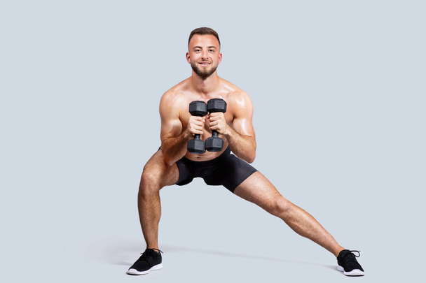 Smiling sweat strong muscular young european man with beard, naked torso make exercises with dumbbells, isolated on gray studio background. Strength workout, sport, body care - Photo, Image