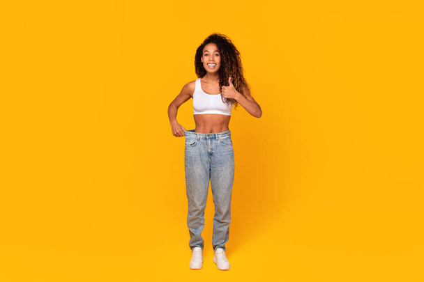 Hymyilevä musta nainen näyttää onnistuneen painonpudotuksen tuloksen peukalot ylöspäin hyväksyvä ruokavalio, yllään vanhat ylisuuret farkut, jotka seisovat keltaisella taustalla. Laihdutus ja laihduttaminen motivaatio - Valokuva, kuva
