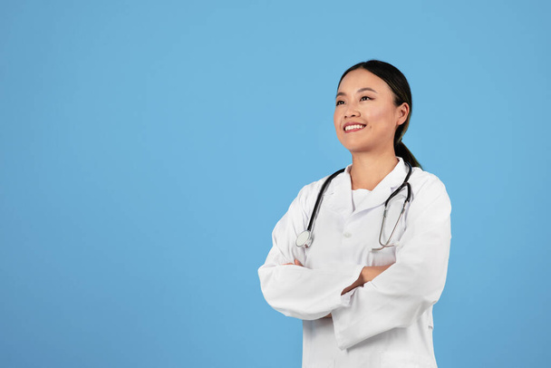 Ιατρικές Υπηρεσίες. Αυτοπεποίθηση ασιατική γιατρός γυναίκα σε ομοιόμορφη στέκεται με διπλωμένα χέρια πάνω από μπλε φόντο στούντιο, γιατρός κυρία φορώντας λευκό παλτό και στηθοσκόπιο κοιτάζοντας μακριά και χαμογελώντας, αντίγραφο χώρο - Φωτογραφία, εικόνα